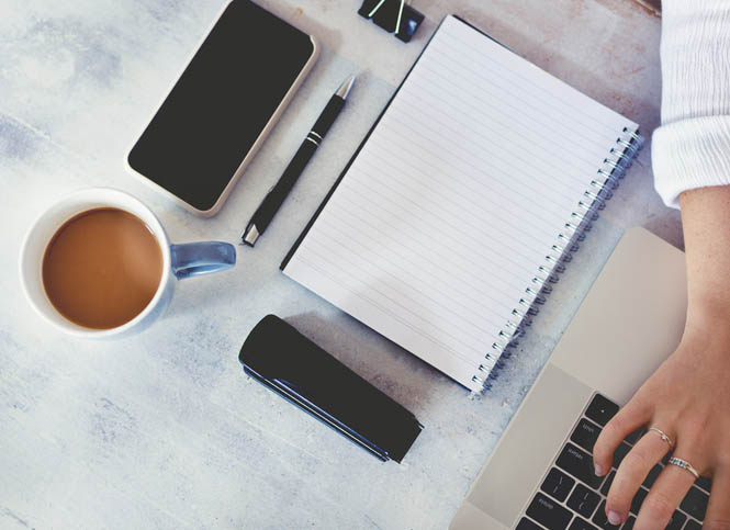 Top view of woman using laptop while sitting at the table with laptop, mobile phone, blank note pad and pen.